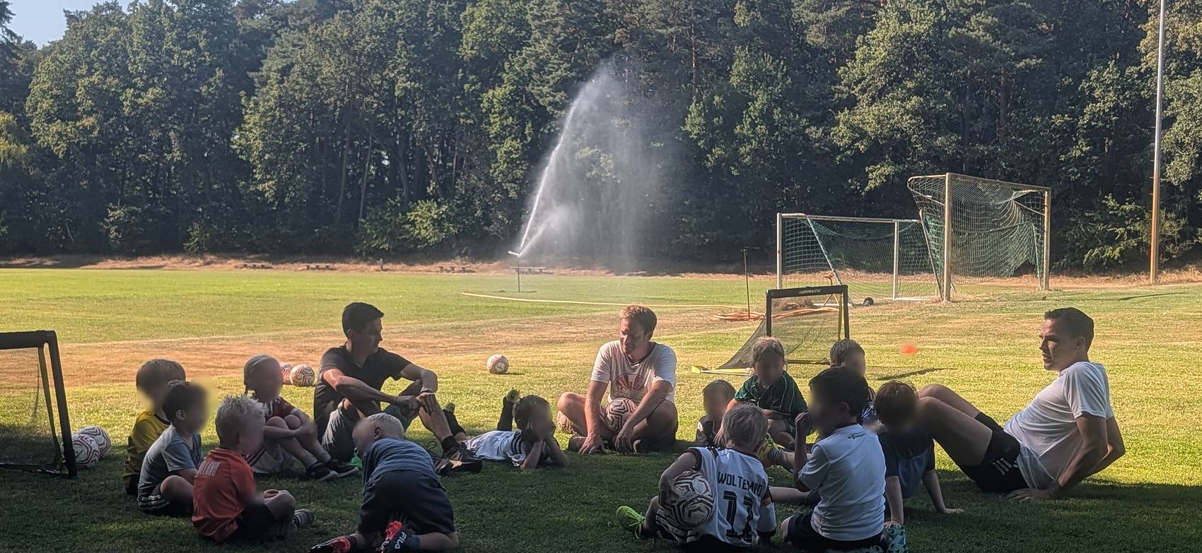 Kindergruppe sitzt nach dem Training im Kreis auf dem Sportplatz, im Hintergrund die Beregnungsanlage